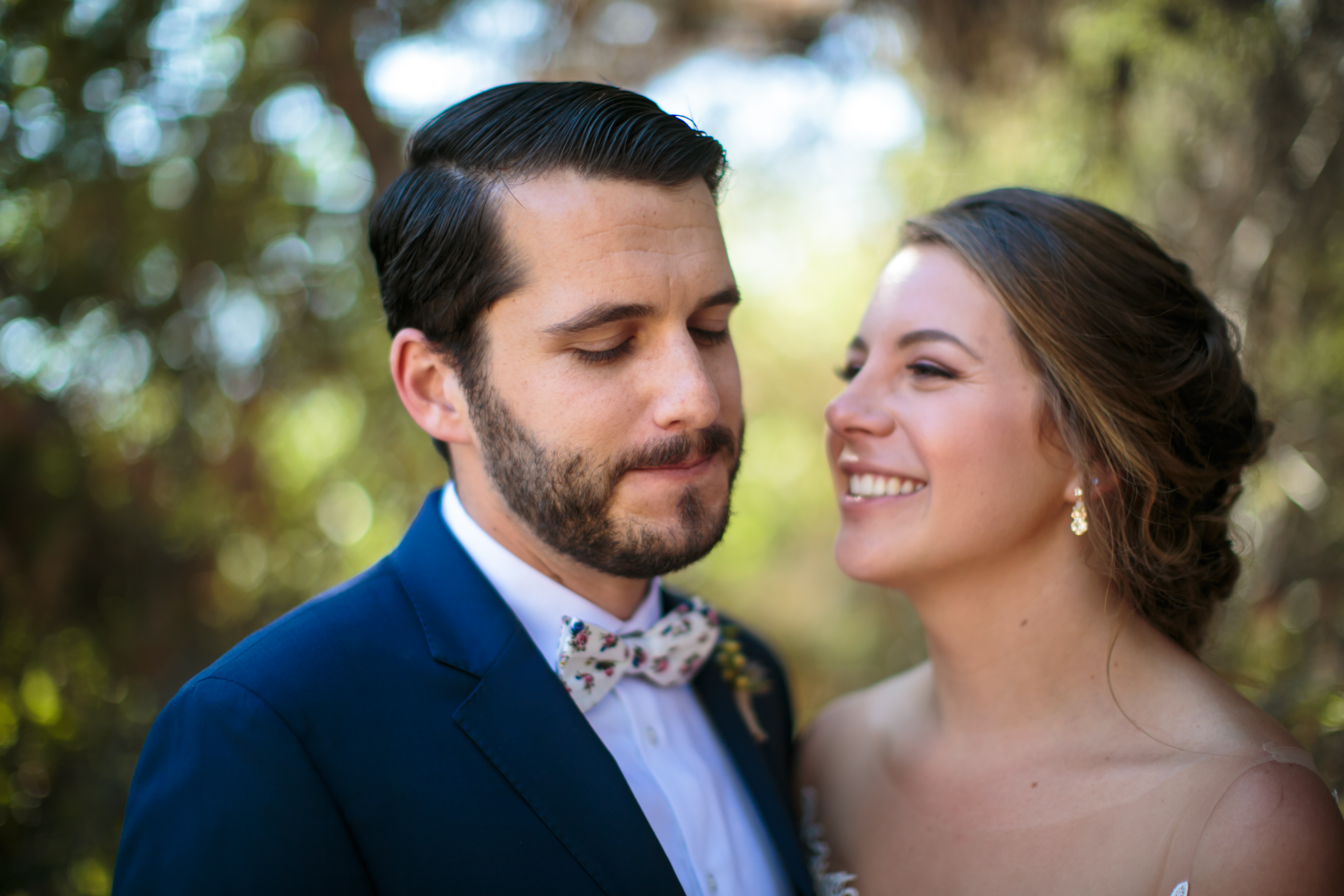 The groom wears a pink and blue floral tie with a blue suit jacket. They are standing in front of some backlit trees and she is smiling at him.