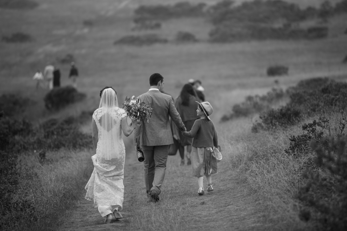 The bride holds her gown in one hand and bouquet in the other. The groom holds the flower girl's hand and a bottle of champagne as they all walk away down a hill side. The girl is wearing a hat and looking up at the groom. It is in black and white.