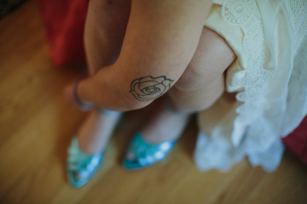 A rose tattoo is on the elbow of the bride as she buckles her blue wedding shoes.