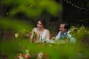 Abstract and out of focus greenery and pink flowers frame the bride and groom sitting and laughing at their sweetheart table.