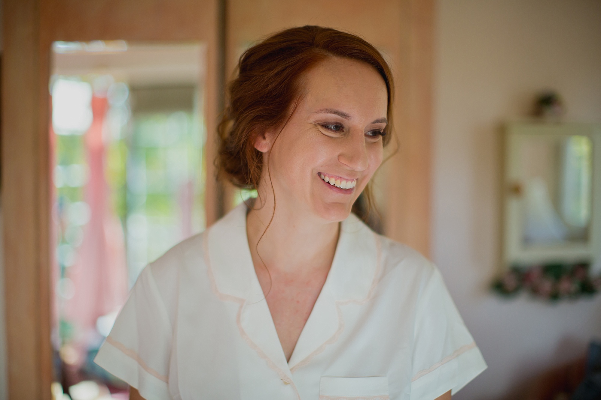 The bride smiles in her white button up shirt and her out of focus blush gown can be seen hanging in the doorway behind her.
