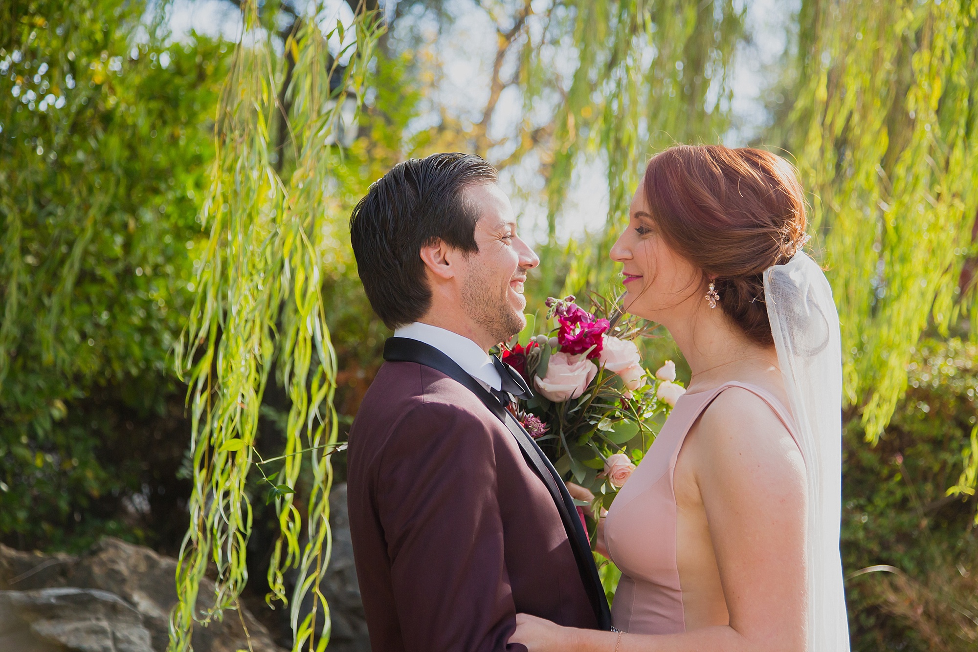 The bride and groom smile at each other while standing under a willow tree.