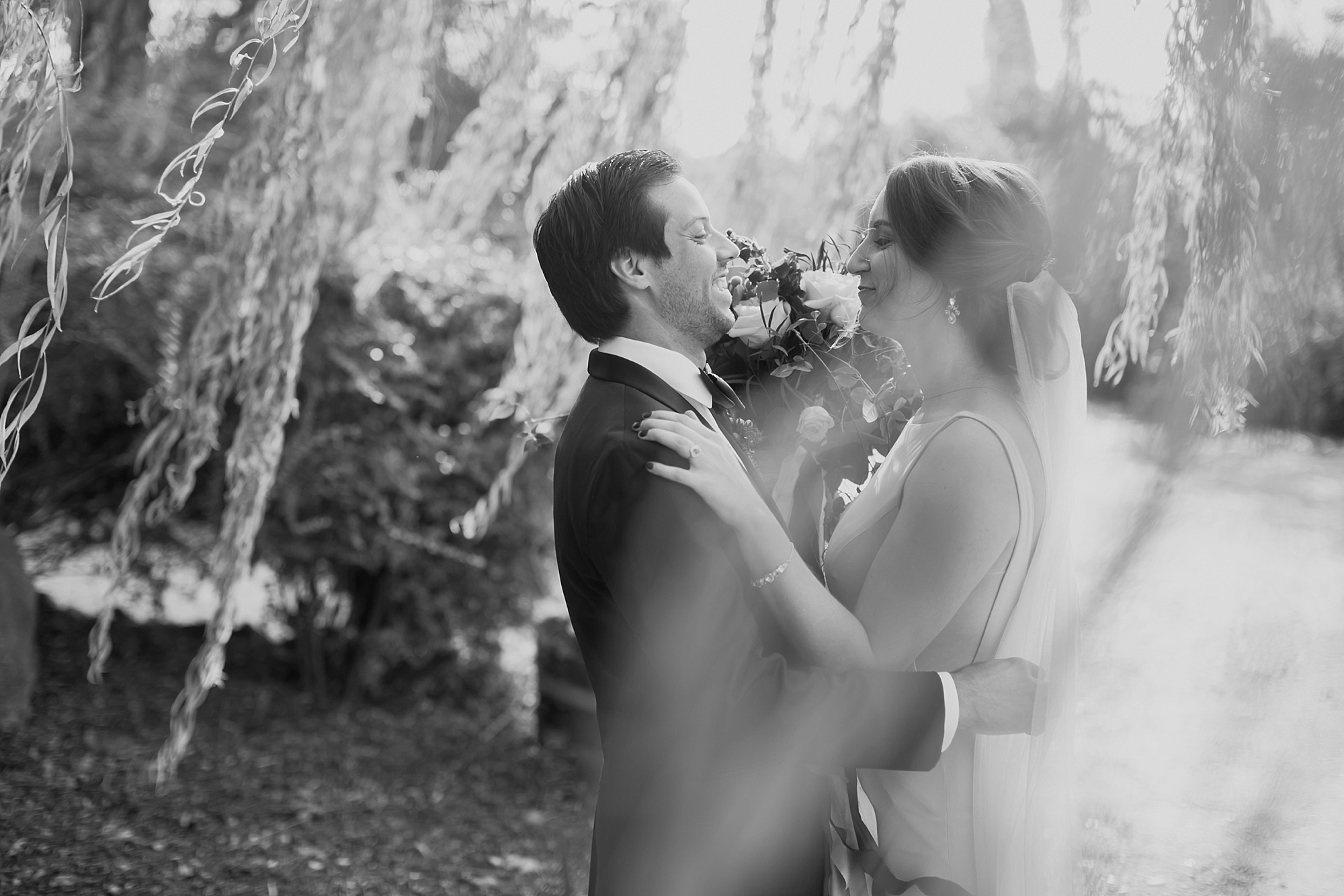 A black and white images of the bride and groom during their first look, surrounded by willow tree leaves.