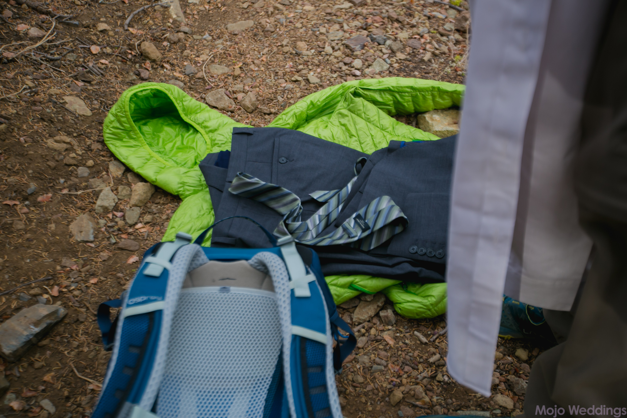 A bright green hiking jacket lays on the ground with wedding pants and tie laying on top of it.