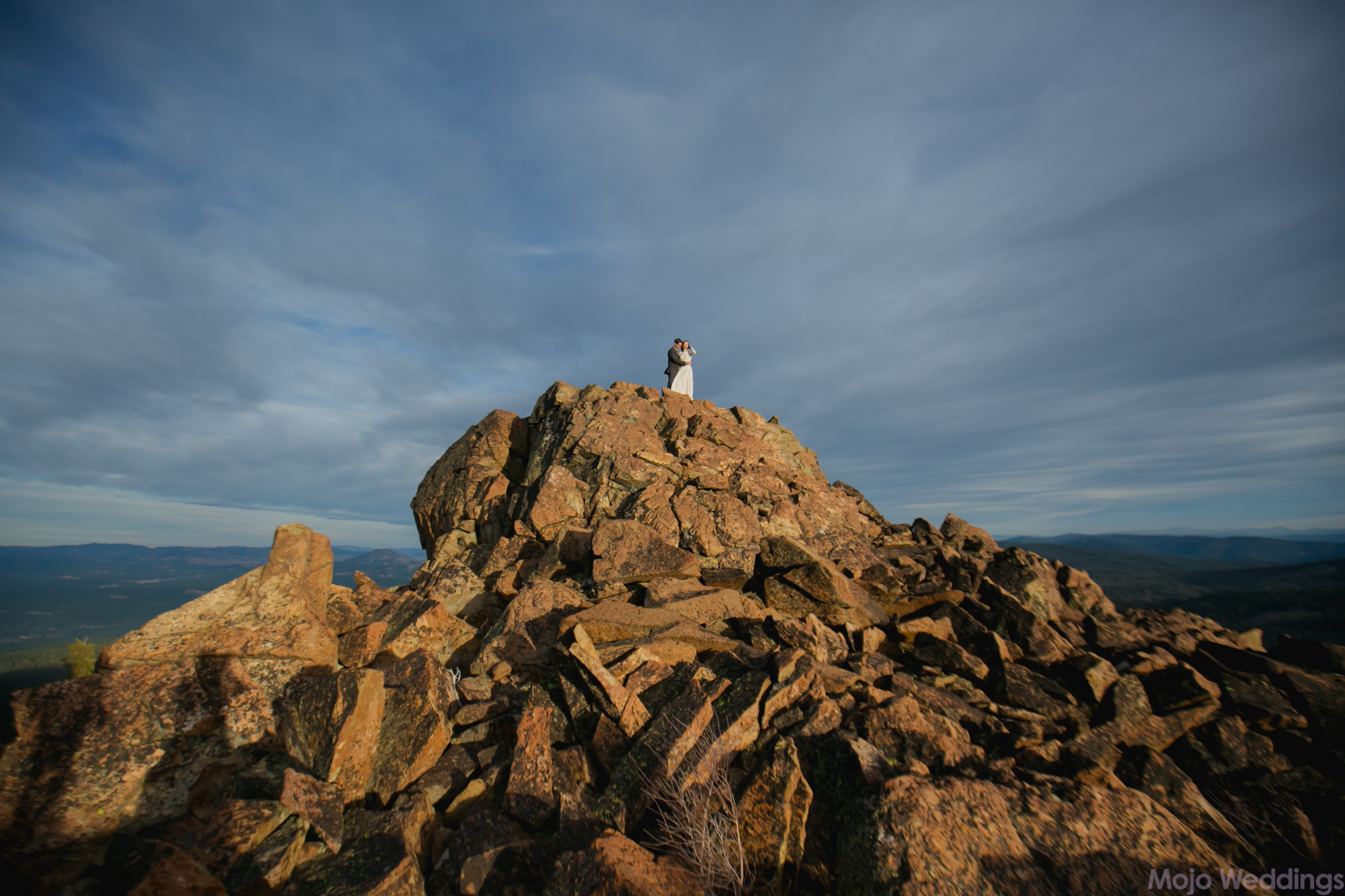 The bride and groom are small on top of a glowing orange rocky mountaintop with dark blue cloudy skies behind them.