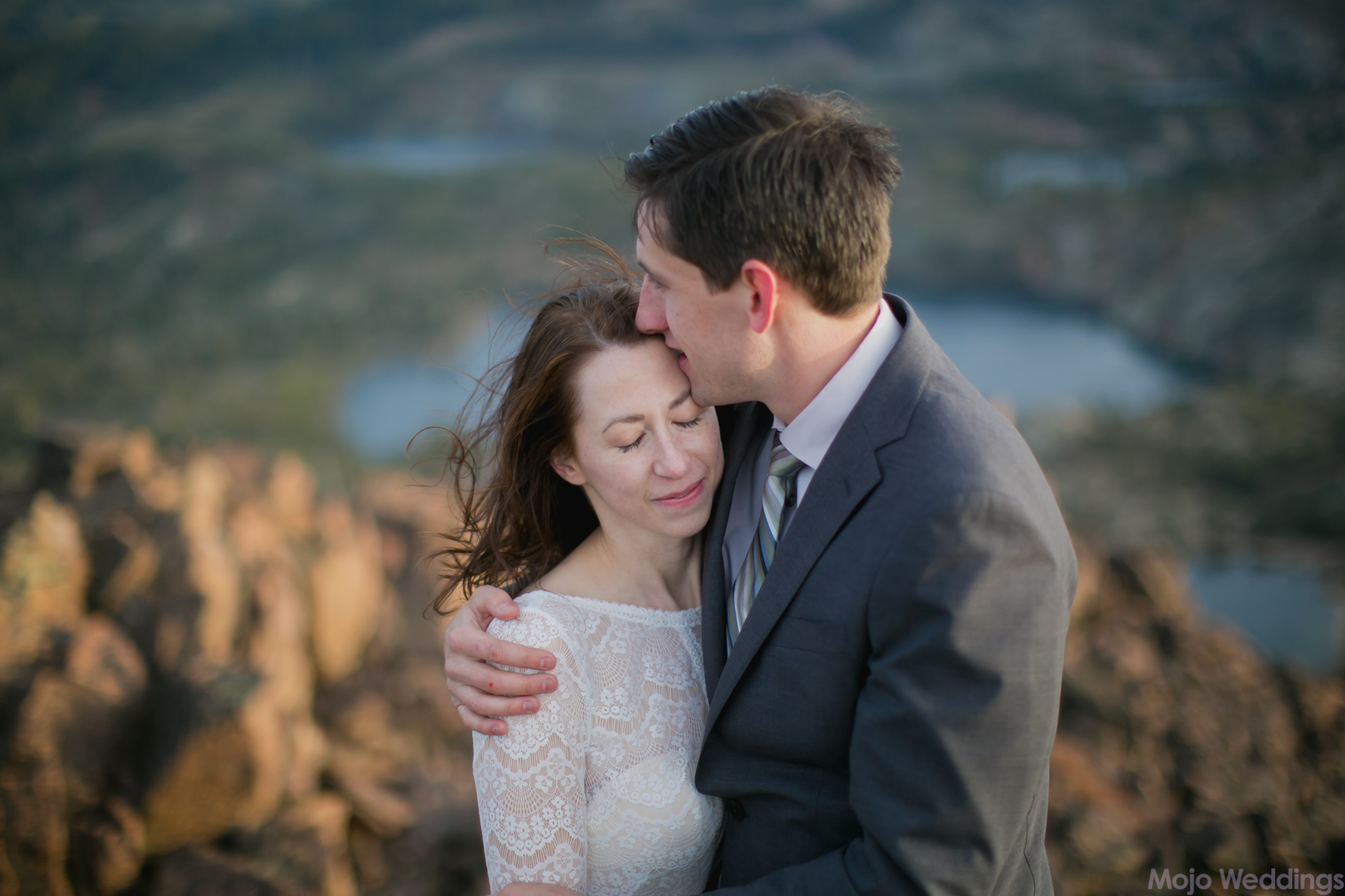 He kisses her forehead with a blurred out background of mountaintop and lakes.